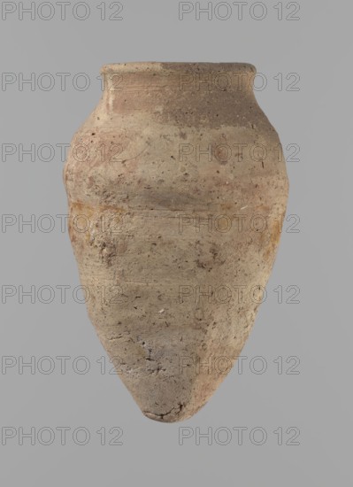 Vessel with hieratic inscription. Conical clay vessel tapering toward the bottom, irregular, widest approx. 3/4 of the height. Narrow, slightly sloping shoulders; short, wide neck; very gently rounded, slightly flaring rim; pointed bottom. Handcrafted; upper part shoulders, neck, and rim turned. Smooth surface, externally probably covered with an engobe. Made of Nile clay NB2 Vienna System. Remains of a hieratic inscription in black on the lower part of the belly. Complete, well preserved. Comparative parallels cited; dated to the Old Kingdom, ca. 2686–2181 BC, find from Edfu. Dimensions: height 12.9 cm; rim diameter 5.5 cm; body diameter 7.8 cm., An ancient-looking clay vessel with a rounded shoulder and pointed base is shown against a plain gray background; the surface of the vessel is rough and weathered with an uneven texture, showing a mix of beige, tan, brown, and muted reddish tones, with some darker brown speckles and lighter patches, and small pits and irregularities across it...