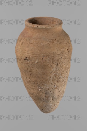 Vessel with hieratic inscription, Egyptian pottery, conical clay vessel tapering toward the bottom, irregular, widest approx. 3/4 of the height. Narrow, only slightly sloping shoulders; short, wide neck; very gently rounded, slightly flaring rim; pointed bottom. Handcrafted; upper part shoulders, neck, rim turned. Smooth surface, exterior probably covered with engobe. In the widest part three shallow horizontal lines over ~0.8 cm. Made of Nile clay NB2 Vienna System. Remains of a hieratic inscription in black sht on lower body. Vessel complete, well-preserved. Comparative parallels cited; earlier dated to the 4th–5th Dynasty / Old Kingdom, ca. 2686–2181 BC. Findspot: Edfu. Dimensions: height 14.4 cm; rim diameter 5.4 cm; body diameter 8.4 cm., A weathered clay jar with a rounded rim and a tapered pointed base fills the frame; its surface is rough and pitted with irregular small holes and encrustations, showing varied earthy tones of brown, tan, and beige with occasional lighter whitish...