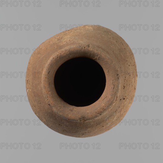 Vessel with hieratic inscription. Conical clay vessel tapering toward the bottom; irregular, widest at approx. 3/4 of the height. Narrow, only slightly sloping shoulders; short, wide neck; very gently rounded, slightly flaring rim; pointed bottom. Handcrafted: upper part shoulders, neck, rim turned. Smooth surface, externally probably covered with engobe. In the widest part three shallow horizontal lines placed over a space of approx. 0.8 cm. Made of Nile clay NB2 Vienna System. In the lower part of the body are remains of a hieratic inscription in black sht. Vessel complete, well-preserved. Comparative parallels cited; earlier dated to the Old Kingdom ca. 2686–ca. 2181 BC, find site Edfu. Dimensions: height 14.4 cm; rim diameter 5.4 cm; body diameter 8.4 cm., A top-down view of a round, open-mouthed clay vessel with a rough, textured surface in shades of brown and tan set against a uniform light gray background, showing the deep dark interior and the uneven rim with small pits and var...