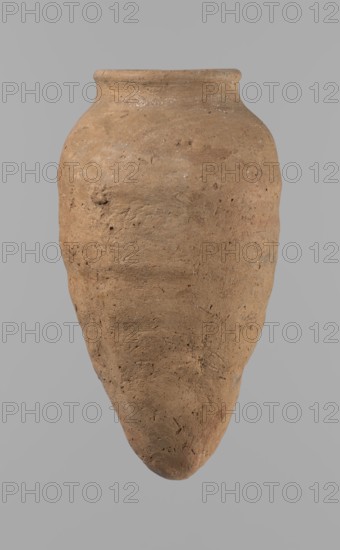 Vessel with hieratic inscription. Clay ceramic hand-made vessel, turned in the upper part neck, spout. Conical shape tapering toward a pointed bottom, irregular, widest at approximately 3/4 of the height. Narrow, slightly sloping shoulders; short, wide neck; rounded, slightly flaring spout. Smooth surface, light red color. Made of Nile clay NB2 Vienna System. Remains of a hieratic inscription in black on the lower part of the belly. Quite well preserved; rim slightly damaged. Old Kingdom, ca. 2686–2181 BC. Height 15.7 cm; diameter rim 5.7 cm; diameter belly 8.5 cm. Comparative parallels cited from Petrie, Reisner, Brunton, Mace, Keiser and others., A large rounded ceramic vessel with a tapered pointed base and a short flared neck, shown against a plain light gray background; the vessel surface is textured and uneven with visible small pits and rough patches, and its color is predominantly earthy brown with subtle variations of tan and ochre and tiny specks of darker brown.
