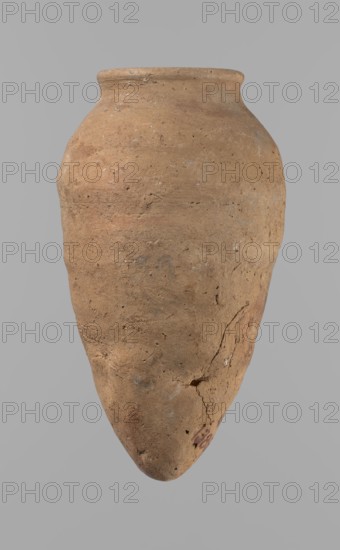 Vessel with hieratic inscription. Hand-made clay vessel, turned in the upper part neck, spout. Conical shape tapering towards the bottom, irregular; widest approx. 3/4 of the height. Narrow, slightly sloping shoulders; short, wide neck; rounded, slightly flaring spout; pointed bottom. Smooth surface. Color light red. Made of Nile clay NB2 Vienna System. Remains of a hieratic inscription in black on the lower part of the belly. Fairly well preserved; rim slightly damaged. Old Kingdom ca. 2686–2181 BC, Edfu. Height 15.7 cm; rim diameter 5.7 cm; belly diameter 8.5 cm., A large earthenware vessel with an elongated, tapered body and a rounded shoulder, shown against a plain gray background; the surface is rough and textured with visible pits, scratches, and small chips, displaying a mix of tan, light brown, beige, and faint reddish-brown tones, with a few areas of darker brown and small pale grayish spots, and a noticeable crack and some surface irregularities near the lower portion.