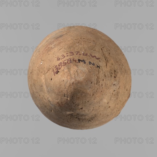 Vessel with hieratic inscription. Clay vessel, hand-made and turned in the upper part neck, spout. Conical shape tapering toward a pointed bottom; irregular, widest at approximately 3/4 of the height. Narrow, slightly sloping shoulders; short, wide neck; rounded, slightly flaring spout. Smooth surface; light red color. Made of Nile clay NB2 Vienna System. Remains of a hieratic inscription in black on the lower part of the belly. Rim slightly damaged; otherwise quite well preserved. Old Kingdom, Edfu. Height 15.7 cm; rim diameter 5.7 cm; belly diameter 8.5 cm. Comparative parallels cited; earlier comparisons date similar types to ca. 2686–2181 BC 4th–5th Dynasty., A round, dome-shaped object with an uneven, textured surface in shades of brown and tan sits against a solid light gray background; the object shows areas of darker brown and subtle reddish-brown variations, small pits and rough patches, and a few darker speckles and smudges.