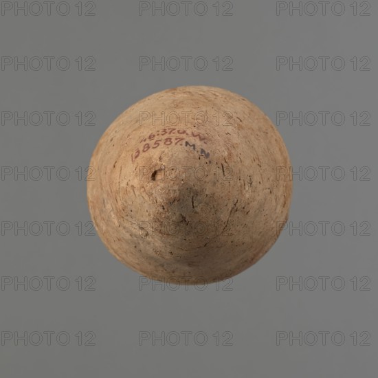 Vessel with hieratic inscription. Conical Nile-clay NB2 pottery vessel, tapering to a pointed bottom; irregular form, widest at roughly 3/4 of the height. Narrow, slightly sloping shoulders; short, wide neck; very gently rounded, slightly flaring rim. Handcrafted, with shoulders, neck, and rim turned. Smooth surface with probable engobe exterior. Remains of a hieratic inscription in black on the lower belly. Good preservation with slight rim damage. Old Kingdom, ca. 2686–2181 BCE. Height 14.8 cm; rim diameter 5.8 cm; belly diameter 8.8 cm. Analogous examples cited in comparative literature; typologically related to vessels dated to the 4th–5th dynasties., A round, cone-topped object with a rough, pitted surface in shades of tan and light brown sits against a uniform medium gray background; the object shows small darker brown speckles and irregular markings, a few shallow pits and surface cracks, and areas of slightly darker brown discoloration.