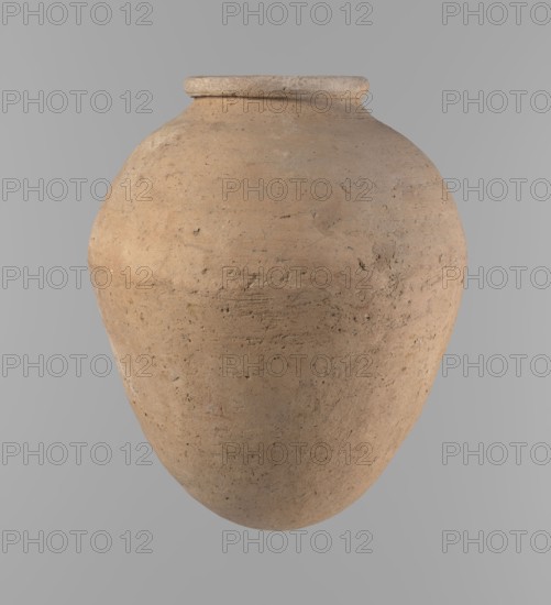 Vessel, Egyptian clay vessel of ovoid, slightly downward-stretched shape, widest at about 2/3 of the height; broad shoulders, very short wide neck, roller-shaped rim oval in cross-section and slightly flaring, rounded bottom. Handcrafted from two parts joined at about 2/3 of the height, turned in the upper part. Surface leveled; traces of a white substance on outer surface and rim in places. Clay with significant organic admixture Viennese system. Color beige. Complete, well-preserved. Late Old Kingdom ca. 2686–ca. 2181 BC. From Edfu. Height 24.9 cm; rim diameter 10.2 cm; body diameter 20.7 cm. References: Rzeuska, Saqqara II; Rzeuska, Chronological overview of pottery from Asyut., A large rounded clay vessel with a narrow rim, showing a rough textured surface with small pits and irregularities; the vessel is mainly tan and light brown with areas of darker brown and subtle grayish tones, set against a smooth light gray background.