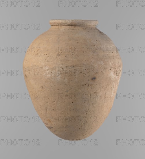Vessel, Egyptian ceramic ovoid vessel, clay with significant organic admixture Viennese System. Handcrafted from two parts joined at about 2/3 of the height, upper part turned. Ovoid, slightly downward-stretched shape, widest at ~2/3 height; broad shoulders, very short wide neck; roller-shaped rim oval in cross-section and slightly flaring; rounded bottom. Surface leveled; traces of a white substance on outer surface and rim. Beige color; complete, well-preserved. Late Old Kingdom ca. 2686–2181 BC. From Edfu. Height 24.9 cm; rim diameter 10.2 cm; body diameter 20.7 cm., A large rounded clay jar with a short, slightly raised rim sits against a plain neutral background; the jar's surface shows a rough, textured finish with small pits and wear marks, and its overall color is a warm beige with patches of lighter tan and occasional darker brown and grayish spots.