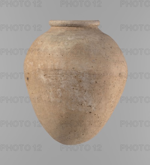 Vessel, Clay ovoid vessel with slightly downward-stretched shape, widest at about 2/3 of the height. Broad shoulders, very short wide neck; roller-shaped rim oval in cross-section, slightly flaring. Rounded bottom. Clay with significant organic admixture Viennese System. Handcrafted from two parts joined at about 2/3 height; upper part turned. Surface leveled; traces of a white substance on outer surface and rim. Beige color. Complete, well-preserved. Late Old Kingdom ca. 2686–2181 BC. From Edfu; height 24.9 cm, rim diameter 10.2 cm, body diameter 20.7 cm., A large rounded clay vessel centered against a plain light gray background, showing an aged textured surface with earthy tones of beige, tan, light brown, and pale gray, a slightly flared rim at the top, a bulbous body tapering toward the bottom, and visible surface irregularities such as small pits, scratches, and mottled patches.