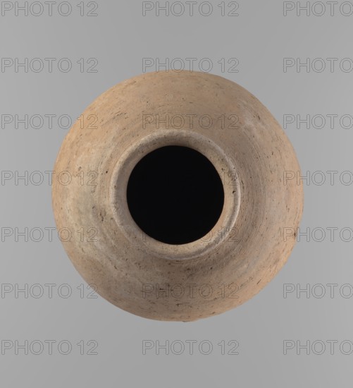 Vessel, Egyptian ceramic clay vessel of ovoid, slightly downward-stretched shape, widest at about 2/3 of the height. Broad shoulders, very short wide neck; roller-shaped rim oval in cross-section, slightly flaring. Rounded bottom. Handcrafted from two parts joined at about 2/3 of the height; upper part turned. Clay with a significant amount of organic admixture Viennese System. Surface leveled; traces of a white substance on outer surface and rim in places. Beige color. Complete, well-preserved. Late Old Kingdom ca. 2686–ca. 2181 BC. From Edfu. Height 24.9 cm; rim diameter 10.2 cm; body diameter 20.7 cm., A round, weathered clay vessel viewed from above with a wide dark opening at its center, showing a rough textured surface in shades of tan, beige, brown, and a few gray speckles, set against a smooth pale gray background.
