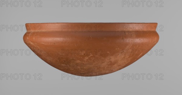 Medum-type bowl, Egyptian ceramic clay vessel, Medum-type bowl with characteristic carination under the rim. Rounded bottom, straight slightly flaring rim, narrow sloping shoulders; rim diameter smaller than shoulder diameter. Surface leveled, covered with engobe and polished. Red color. Vessel cracked and glued; minor clay losses on rim filled with plaster. Old Kingdom ca. 2686–ca. 2181 BC; possibly 6th Dynasty. From Edfu. Height 8.9 cm; rim diameter 21.7 cm; shoulder maximum diameter 22.3 cm., A close-up of a rounded, shallow bowl-shaped object in a terracotta color with a matte surface showing a few scuffs and uneven patches, set against a plain light gray background; the object has a slightly flared rim and a smooth curved profile with a few small dark specks and areas of lighter wear.