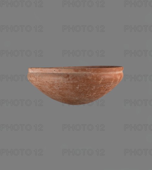 Medum type bowl. Medum type bowl with characteristic carination under the rim. Rounded bottom. Straight rim, slightly flaring. Narrow, sloping shoulders. The diameter of the rim is smaller than the diameter of the vessel's shoulders. Clay probably NB1. Surface leveled and covered with a layer of engobe, then polished. Vessel color - red. Complete vessel, very well preserved. c. 2686–c. 2181 BC; Old Kingdom, Edfu. Vessel, height 4.2 cm, diameter 21.1 cm rim, diameter 21.1 cm shoulders., A single shallow, rounded clay bowl with a slightly raised rim sits centrally against a plain background; the bowl shows a weathered surface with scratches, patches of lighter wear, and small pits, and the visible colors include terracotta, light brown, pale beige, and hints of gray.