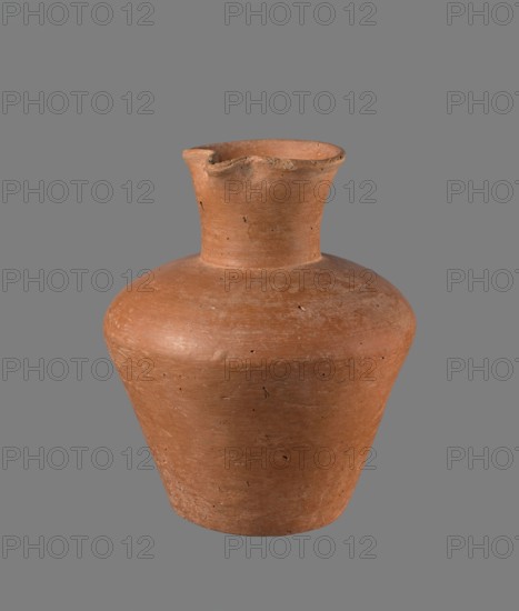 Jug, Egyptian pottery, cylindrical clay vessel with flaring neck, c. 2686–c. 2181 BC Old Kingdom, from Edfu; height 18.6 cm, rim diameter 8.5 cm, body diameter 15.8 cm, bottom diameter 9.1 cm., A clay vessel with an earthy reddish-brown color sits against a flat gray background; the vessel has a rounded shoulder that tapers to a narrower base and rises to a short neck with a flared rim that shows a small chipped area, and the surface displays a matte, slightly rough texture with small pits and darker flecks.