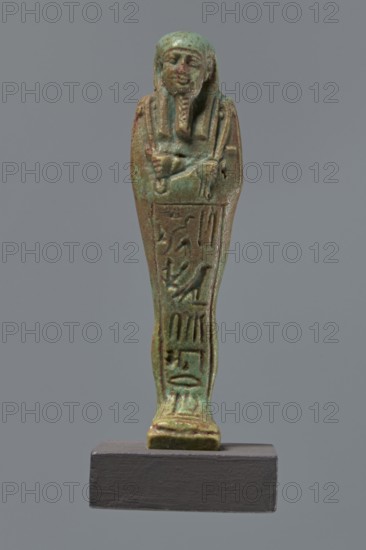 Ushabti Hora, Mummy-shaped figurine shabti, mummy-shaped figurine, standing on a base, back supported by a pillar/pilaster. Arms crossed on the chest, right folded over the left. Hoes in both hands. On the back is a basket, slung on a single string over the left shoulder. On the head is a three-part, smooth wig. Long braided beard. Face with proportional features, mouth set in a slight smile. Inscribed inscription on the front of the legs in one wide column framed with lines: Shd Osiris Hor born of Her-ib, justified by the voice. Ptolemaic Period, Egypt, 332–30 BC. Height 11.1 cm., A small statuette of a standing mummiform figure is shown against a plain grey background, displayed on a dark rectangular base; the figure has a stylized human face with a headdress and a false beard, arms crossed over the chest, and a narrow elongated body with carved relief details along the front. The surface shows a weathered greenish-blue patina with areas of brown and darker green, and a few spots of ...
