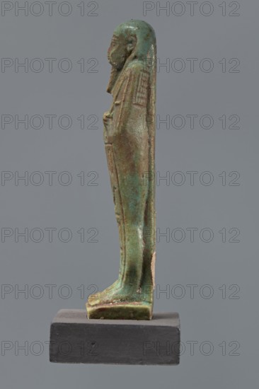 Ushabti Hora, Mummy-shaped figurine, funerary shabti, Ptolemaic Period, 332–30 BC, Egypt. Standing on a base, back supported by a pillar. Arms crossed on the chest, right over left, holding hoes in both hands. Basket on the back, slung on a single string over the left shoulder. Wearing a three-part smooth wig and a long braided beard. Face with proportional features and a slight smile. Inscribed on the front of the legs in one wide column framed with lines: Shd Osiris Hor born of Her-ib, justified by the voice. Height 11.1 cm., A slender statuette of a standing human figure displayed in profile on a dark rectangular base, with a smooth rounded head and elongated body draped in a long garment that reaches the feet; the surface shows a weathered, mottled appearance in shades of green and turquoise with patches of brown and darker green, and traces of a lighter beige material adhered to one side; the base is matte dark gray.
