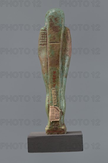 Ushabti Hora, Mummy-shaped figurine shabti, funerary gift, 332–30 BC; Ptolemaic Period, Egypt. Standing on a base, back supported by a pillar/pilaster. Arms crossed on the chest, right folded over the left, holding hoes in both hands. A basket is on the back, slung on a single string over the left shoulder. Wearing a three-part smooth wig and a long braided beard. Face with proportional features, mouth set in a slight smile. Inscribed inscription placed on the front of the legs in one wide column framed with lines: Shd Osiris Hor born of Her-ib, justified by the voice. Height: 11.1 cm., A small statuette viewed from the back standing on a dark base, with a worn surface showing shades of green, turquoise, brown, and hints of reddish-brown; the figure has a rounded head and a long vertical form with carved vertical and horizontal grooves, and a few small fabric patches adhered to the lower back area; the background is a uniform gray.