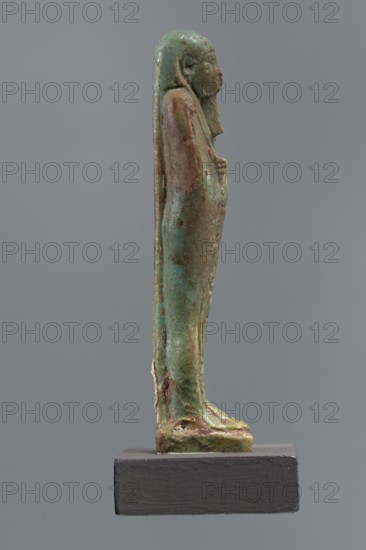 Mummy-shaped shabti of Horus inscribed Shd Osiris Hor born of Her-ib, justified by the voice, Ptolemaic Period, Egypt, 332–30 BC. Standing on a base with back supported by a pillar; arms crossed on the chest right over left holding hoes in both hands; a basket on the back slung over the left shoulder by a single string. Wears a three-part smooth wig and a long braided beard. Face with proportional features and a slight smile. Inscription in one wide column framed with lines on the front of the legs. Height 11.1 cm., A slender standing human figure sculpture shown in profile on a rectangular base, with a smooth elongated form and a head with discernible facial features and long hair or headdress flowing down the back; the surface shows varied green and brown patina with areas of lighter turquoise and darker verdigris, and some rougher brownish spots and worn textures, while the base is a solid dark gray.