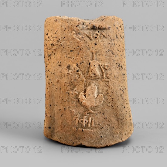 Fragment of an amphora vice behind a stamp, Sinope Asia Minor uncertain, 4th–beginning of 3rd century BC, Pantikapaion, vessel; height 4.1 cm, width 6.3 cm, A single upright cylindrical object with a rough, porous surface in shades of brown and tan, dotted with small dark speckles, resting on a smooth light gray surface against a neutral light gray background; the object shows carved or incised shallow relief shapes on its front face including rounded and angular forms, with some areas showing lighter beige highlights where the surface texture varies, and the top edge appears uneven and slightly chipped.