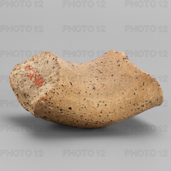 Fragment of an amphora neck with a stamp, Sinope Asia Minor uncertain, 4th–beginning of 3rd century BC, Pantikapaion; vessel; height 4.1 cm, width 6.3 cm, A broken curved fragment of pottery or ceramic rests on a smooth gray surface against a gray background; the object is mainly tan with a speckled pattern of small black flecks and a few darker brown spots, showing rough, uneven edges and a concave inner curve, with one end appearing more jagged and textured while the opposite side is smoother and rounded.