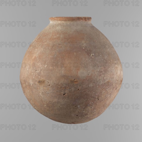 Vessel with an almost spherical belly, widest about 1/3 of the vessel's height. Sloping shoulders. Neck poorly defined – very short and wide. Rounded, slightly flaring rim. Rounded bottom. Made of Nile clay NB. Surface smooth; outer surface finished to about half its height with a knife or similar tool. A rather indistinct cord impression around the widest diameter of the belly. Light red color. Chipped rim and traces of salting in places over the entire surface. Comparative material: Reisner, Mycerinus, fig. 62.3 type II, III, ca. 2686–2181 BC; Old Kingdom, Edfu, vessel, height 24.9 cm, diameter 10.3 cm rim, diameter 24.2 cm body., A large rounded clay vessel with a narrow rim, showing a weathered, textured surface in shades of brown, reddish-brown, tan, and gray, with small pits, scratches, and uneven patches across its curved body, set against a smooth light gray background.