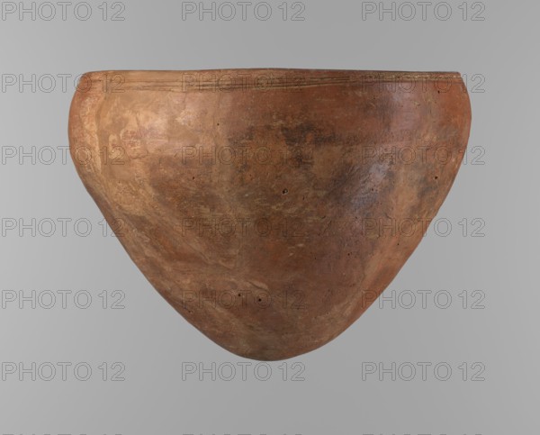 Bowl, Egyptian pottery; clay vessel, large conical vessel with widest point about 4/5 of the height, walls narrowing toward center, rounded bottom, straight slightly tapering spout. Two parallel horizontal shallow lines just below the rim. Made of Nile clay NB2 Vienna System. Surface leveled and covered with a layer of red engobe; overall dark red color. Reconstructed from original fragments with losses, including significant rim fragments, filled with plaster. Dated end of predynastic ST.P / beginning of 1st Old Kingdom? References to parallels: F. Petrie Medum and A. Fakhry monuments of Sneferu at Dahshur. Origin: Edfu; Old Kingdom, c. 2686–2181 BC. Vessel height 21.4 cm; diameter 28.5 cm rim, 30.5 cm shoulders., A large rounded clay vessel with an inverted bulbous shape fills the frame, showing a textured surface with earthy brown, reddish-brown, and terracotta tones along with subtle patches of darker brown and muted orange; the rim has a few shallow incised lines and the body disp...