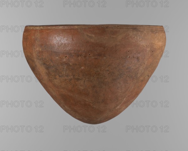 Bowl, Egyptian pottery clay vessel, large conical vessel with widest about 4/5 of the height, walls narrowing toward the center, rounded bottom, straight slightly tapering spout. Just below the rim are two parallel horizontal shallow lines. Made of Nile clay NB2 Vienna System. Surface leveled and covered with red engobe; vessel color dark red. Reconstructed from original fragments with losses, including significant rim fragments filled with plaster. Dated to end of ST.P beginning of 1st OP? Old Kingdom, Edfu; c. 2686–2181 BC. Height 21.4 cm, rim dia. 28.5 cm, shoulder dia. 30.5 cm. References: F. Petrie, Medum, pl. XXXI, fig. 3; A. Fakhry, The Monuments of Sneferu at Dahshur, p. 116, figs. 7-5, c., A large rounded clay pot with a wide opening and a gently tapered bottom fills the frame, showing an uneven textured surface with small pits and patches of wear; the pot displays shades of reddish brown, terracotta, muted orange, and hints of darker brown and gray, and it sits against a smoo...