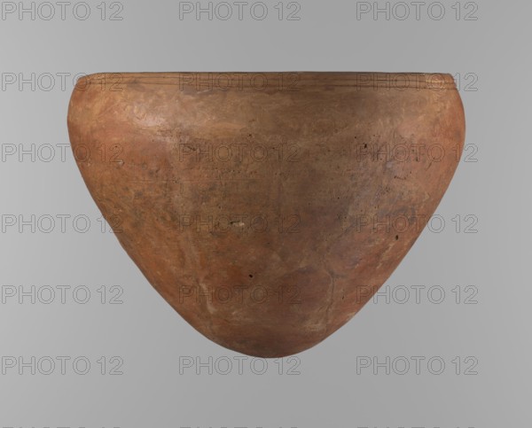 Bowl, Large conical Egyptian pottery vessel, clay; wide about 4/5 of the height with walls narrowing toward center, rounded bottom, straight slightly tapering spout. Two parallel horizontal shallow lines just below the rim. Made of Nile clay NB2 Vienna System. Surface leveled and covered with red engobe; dark red color. Reconstructed from original fragments with losses, significant rim fragments filled with plaster. Dated end of ST.P / beginning of 1st OP Old Kingdom, c. 2686–2181 BC. From Edfu. Vessel height 21.4 cm; rim diameter 28.5 cm; shoulder diameter 30.5 cm., A large rounded clay vessel with a wide rim is centered against a plain gray background; the surface of the vessel shows a weathered, uneven texture with a mix of earthy red, brown, tan, and subtle orange tones, along with darker brown spots and small pits, and faint lighter streaks and patches across its curved body.