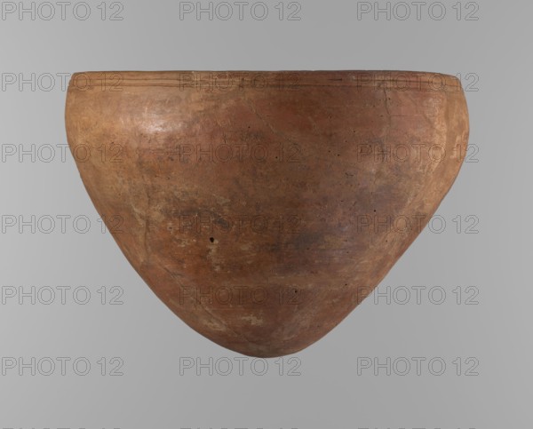 Bowl, Egyptian pottery, clay vessel; large conical vessel with widest point about 4/5 of the height, walls narrowing toward center. Rounded bottom; straight slightly tapering spout. Just below the rim two parallel horizontal shallow lines. Made of Nile clay NB2 Vienna System. Surface leveled and covered with a layer of red engobe; dark red color. Reconstructed from original fragments with losses, including significant rim fragments filled in with plaster. Dating: end of 4th Dynasty / beginning of 5th Dynasty c. 2686–c. 2181 BC, Old Kingdom, from Edfu. Height 21.4 cm; diameter: rim 28.5 cm, shoulders 30.5 cm. References: F. Petrie; A. Fakhry., A large rounded clay vessel with a wide open top and a bulbous, tapered body is centered against a smooth light gray background; the vessel surface shows a weathered, earthy reddish-brown color with areas of lighter tan and darker brown patches, faint surface scratches and small pitting, and subtle variations in texture and sheen across its curved...