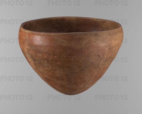 Bowl. Large conical ceramic vessel with rounded bottom and straight slightly tapering spout; widest point about 4/5 of the height where walls begin to narrow toward center. Just below the rim are two parallel horizontal shallow lines. Made of Nile clay NB2 Vienna System. Surface leveled and covered with a layer of red engobe; overall dark red color. Reconstructed from original fragments with losses, including significant rim fragments filled with plaster. Dated end of ST.P / beginning of 1st OP Old Kingdom, c. 2686–2181 BC; provenance Edfu. Height 21.4 cm; diameter 28.5 cm rim, 30.5 cm shoulders. References: F. Petrie, Medum, pl. XXXI, fig. 3; A. Fakhry, The monuments of Sneferu at Dahshur, p. 116, figs. 7-5., A large rounded clay pot with a wide open top fills the frame, showing a textured surface with a weathered, earthy appearance in shades of brown, reddish-brown, and terracotta, and a slightly darker interior; the rim is slightly raised with a faint banded detail, and the body dis...