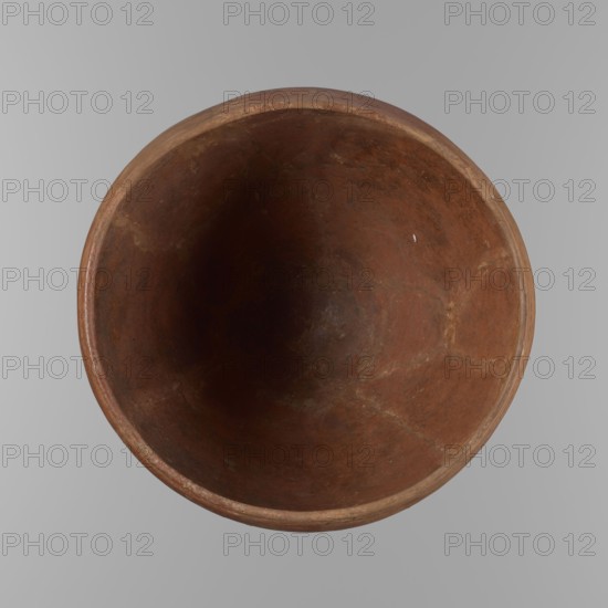 Bowl. Large conical clay vessel, rounded bottom, straight slightly tapering spout; widest about 4/5 of the height where walls begin to narrow toward the center. Just below the rim are two parallel horizontal shallow lines. Made of Nile clay NB2 Vienna System. Surface leveled and covered with a layer of red engobe; vessel color dark red. Reconstructed from original fragments with losses, including significant fragments of the rim, filled in with plaster. Dated end of ST.P beginning of 1st OP? Old Kingdom, c. 2686–2181 BC. Height 21.4 cm; diameter 28.5 cm rim, 30.5 cm shoulders. References: F. Petrie, Medum, pl. XXXI, fig. 3; A. Fakhry, The monuments of Sneferu at Dahshur, p.116, figs. 7-5., A top-down view of a round, hollow clay vessel with a smooth rim and an uneven interior surface showing a few hairline cracks and variations in texture, the vessel displaying shades of brown and reddish-brown with some darker brown shadowing toward the center, set against a light gray background.