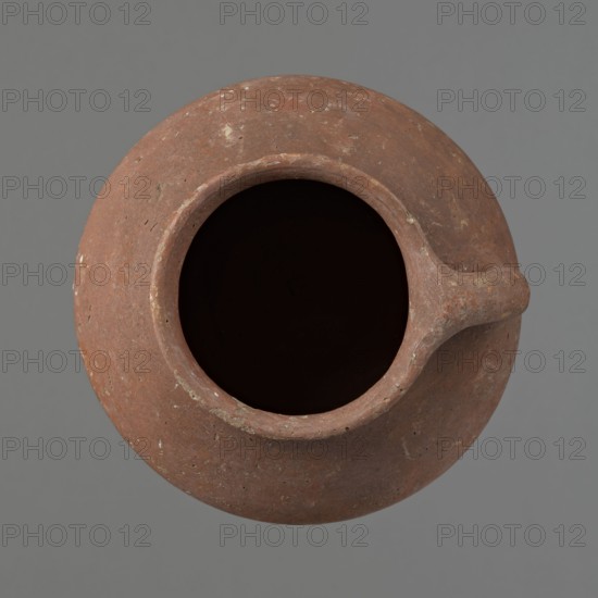 Jug. Small clay jug with one handle; wide, sloping shoulders, short neck widening toward a straight, slightly flaring spout. Handle attaches slightly above the vessel’s widest diameter and extends about 1 cm above the spout line; circular in cross-section below, slightly flattened above. Slightly convex bottom. Made of Nile clay; exterior and rim/neck interior coated with a thick red slip; overall red-brown color. Complete and fairly well preserved; slip worn in places. Old Kingdom, ca. 2686–2181 BC. Dimensions: height 12.7 cm; rim diameter 8.4 cm; body diameter 13.5 cm; bottom diameter 9.5 cm., A top-down view of a round clay vessel with a wide open mouth and a small protruding spout, showing a weathered surface in shades of reddish-brown and terra cotta with lighter flecks and patches, the interior appearing very dark, set against a flat gray background.