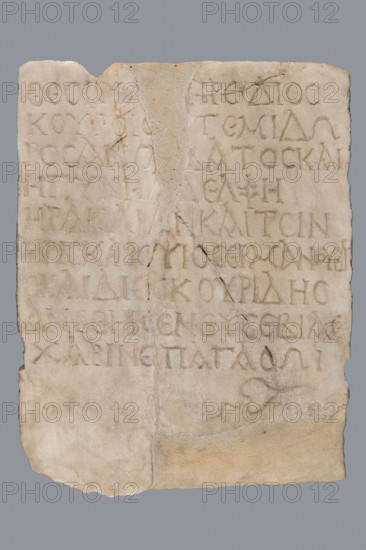 Greek dedicatory inscription, unknown author, tombstone inscription: To the Savior Gods Dioskuros Artemidoros son of Anubatos and his wife-sister Heraklia called also Tsinyris and son Hermanubis called also Dioscorides — gave the grace of piety for happiness hedera. 1st half of the 1st century BC, Egypt. Greek inscription. Height 40 cm, width 30 cm, depth 2 cm., A roughly rectangular carved stone slab with an uneven top edge and a chipped bottom corner, showing a pale off-white and beige surface with areas of light brown discoloration and darker tan patches; the slab has a vertical crack and a visible repair seam with a slightly different beige tone, and a few darker hairline fractures radiating across the surface; the stone bears a group of shallow incised marks forming rows across most of the face, with some marks more worn and faint than others; there is a small carved decorative motif near the lower right area with subtle shadowing; the background behind the slab is a uniform mediu...