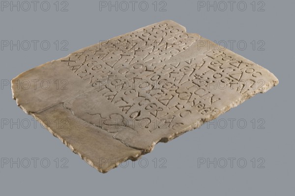 Greek dedicatory inscription, tombstone inscription: To the Savior Gods Dioskuros, Artemidoros son of Anubatos and his wife-sister Heraklia called also Tsinyris and son Hermanubis called also Dioscorides gave the grace of piety for happiness hedera. 1st half of the 1st century BC, Egypt. Greek inscription. Height 40 cm, width 30 cm, depth 2 cm., A flat, irregular stone slab rests against a plain gray background, showing a worn, pale beige surface with rough, chipped edges and a few smoother, slightly darker patches; the slab is inscribed with shallow incised shapes arranged in rows across its surface, and there are faint surface cracks and small pits visible, with overall colors of beige, off-white, light brown, and gray.