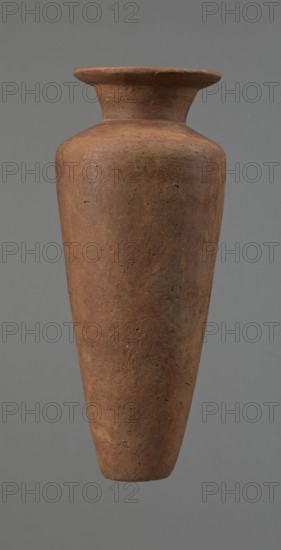 Vessel, Egyptian pottery, Type HS vessel, conical slender shape, widest at about 3/4 of the height. Shoulders wide, almost horizontal, slightly sloping. Neck wide, short, funnel-shaped in lower part, widening towards the rim from about half its height. Rim wide, flat on top, slightly rounded at bottom, slightly asymmetrical. Bottom flat, slightly asymmetrical. Neck made separately and glued to the shoulders. Surface even; lower part finished to approx. 3/4 of height with a knife or similar tool. Outer surface and inner surface of rim and neck covered with dark red engobe and polished. Color dark red. Reconstructed from many original fragments; gaps filled with plaster. c. 2686–c. 2181 BC Old Kingdom, Edfu. Vessel; height 24.8 cm, diameter rim 8.9 cm, diameter shoulders 10.3 cm, diameter bottom 4.1 cm., A tall, narrow vase with a flared rim and a tapered body suspended against a plain background; the surface shows a rough, textured finish with visible small pits and irregularities. The ...