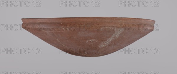 Medum-type bowl, Egyptian pottery, Medum-type bowl with characteristic carination under the rim. Rounded bottom. Straight, slightly flaring rim. Narrow, sloping shoulders. The diameter of the rim equals the diameter of the vessel's shoulders. Clay probably NB1. The surface was leveled and covered with a layer of engobe, then polished. Vessel color red. Vessel glued together from original fragments; minor losses on the rim. c. 2686–c. 2181 BC Old Kingdom, Edfu. Vessel; height 6.9 cm; diameter 24.5 cm rim, 24.9 cm shoulders., A shallow, rounded ceramic bowl shown upside down against a pale gray background, with an overall reddish-brown surface and a slightly raised rim that has visible wear and small chips; the exterior shows lighter, whitish streaks and smudges, a few hairline cracks, and subtle variations in the reddish-brown tone giving a textured, weathered appearance.