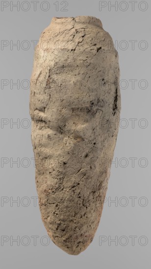 Vessel, Egyptian pottery vessel with a conical belly widest in its upper part approx. 4/5 of the height. Rounded bottom. Irregularly marked shoulders—clearly sloping on one side, short and almost horizontal on the opposite. Straight, slightly tapering rim. Hand-made of not well-worked Nile clay with numerous organic inclusions and limestone particles visible on the surface. Uneven, brown surface with numerous depressions and cracks resulting from poor clay kneading. Ca. 2686–2181 BC Old Kingdom, from Edfu; height 38 cm, diameter 11.7 cm rim, diameter 15.7 cm body., A roughly elongated, irregularly shaped object resembling a weathered stone or clay form fills the image against a plain light gray background; its surface is textured with cracks, pits, and rough patches, showing uneven contours and faint depressions that suggest subtle facial-like features, with a rounded top tapering to a narrower bottom. The visible colors are mainly beige and tan with areas of light brown and small hint...