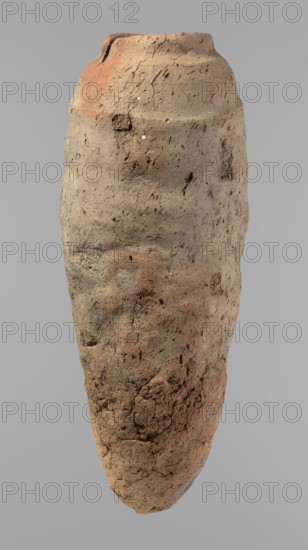 Vessel, Egyptian pottery, hand-made clay vessel with a conical belly widest in its upper part approx. 4/5 of the vessel's height, rounded bottom, irregularly marked shoulders clearly sloping on one side, short and almost horizontal on the opposite, straight slightly tapering rim. Made of not very well-worked Nile clay with numerous organic inclusions and limestone particles visible on the uneven surface; carelessly made with many uneven depressions and surface cracks resulting from poor clay kneading. Brown color. Ca. 2686–2181 BC Old Kingdom, from Edfu. Vessel; height 38 cm, rim diameter 11.7 cm, body diameter 15.7 cm., An upright, elongated clay vessel with a rough, weathered surface, showing a mix of beige, tan, light brown, and patches of reddish-brown hues; the vessel has an irregular, slightly tapered shape with a rounded base and a narrower, uneven rim at the top, and its exterior displays cracks, pits, and textured marks along with a few small, lighter spots against a plain gra...