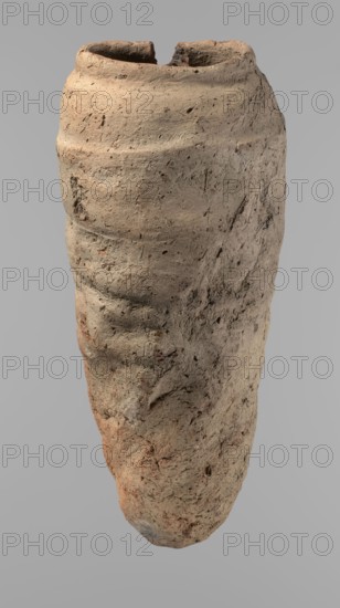 Vessel, Egyptian pottery, hand-made clay vessel with a conical belly widest in its upper part approx. 4/5 of the vessel's height, rounded bottom. Shoulders irregularly marked—clearly sloping on one side and short, almost horizontal on the opposite. Straight, slightly tapering rim. Made of Nile clay with numerous organic inclusions and limestone particles visible on the surface. Surface uneven with numerous uneven depressions; brown color. Carelessly made; cracks on the surface result from poor clay kneading. Old Kingdom, ca. 2686–ca. 2181 BC; from Edfu. Vessel height 38 cm; rim diameter 11.7 cm; body diameter 15.7 cm., A single tall, irregularly shaped clay vessel with a rough textured surface in shades of beige, tan, light brown, and hints of reddish-brown, showing a weathered, pitted appearance and uneven ridges; the top rim is broken and reveals a darker, earthier interior, and the body displays small holes, scratches, and areas of darker brown and gray discoloration against a plain...