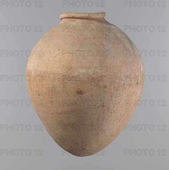 Vessel, Egyptian pottery, hand-made clay vessel of an ovoid, slightly downward-stretched shape, widest at about 2/3 of the height; broad sloping shoulders, very short wide neck, roller-shaped rim oval in cross-section and slightly flaring, rounded bottom. Made from NB2 clay Viennese System with significant organic temper. Constructed from two joined parts join at ~2/3 height, upper part turned. Surface leveled, possibly covered with a layer of engobe; traces of a white substance on outer surface and rim. Beige color. Well preserved. Old Kingdom, ca. 2686–ca. 2181 BC; provenance: Edfu. Vessel; height 34.6 cm, rim diameter 11.6 cm, body diameter 29 cm., A large rounded clay vessel with a narrow rim and an overall weathered, textured surface fills the frame, showing a palette of warm beige, light tan, and subtle reddish-brown tones with patches of pale gray and small darker brown spots, set against a smooth neutral gray background.