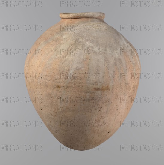 Vessel, Egyptian clay vessel of ovoid, slightly downward-stretched shape, widest at about 2/3 of the height; broad sloping shoulders, very short wide neck; roller-shaped rim oval in cross-section, slightly flaring; rounded bottom. Hand-made from two parts joined at about 2/3 of the height, upper part turned. Surface leveled, possibly covered with a layer of engobe; traces of a white substance in places on the outer surface and on the rim. Clay corresponds to NB2 Viennese system with a significant organic admixture. Colour beige. Well-preserved. Old Kingdom, ca. 2686–ca. 2181 BC; provenance Edfu. Vessel; height 34.6 cm, rim diameter 11.6 cm, body diameter 29 cm., A large rounded ceramic vessel with a narrow rim, showing a weathered, textured surface in shades of beige, tan, light brown, pale gray, and faint peach, with irregular patches and streaks of slightly darker and lighter tones and small surface pits and scratches against a plain neutral gray background.