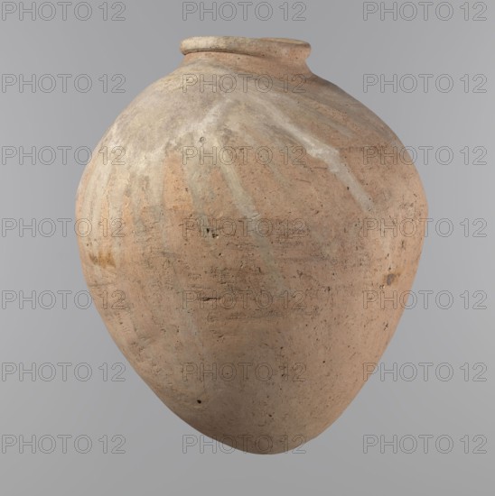 Vessel, Egyptian pottery vessel of ovoid, slightly downward-stretched shape, widest at about 2/3 of the height. Broad, sloping shoulders, very short wide neck. Roller-shaped rim, oval in cross-section, slightly flaring. Rounded bottom. Made of NB2 clay Viennese System with significant organic admixture. Hand-made from two parts joined at about 2/3 of the height, turned in the upper part. Surface leveled possibly covered with a layer of engobe. Traces of a white substance in places on the outer surface of the body and on the rim. Beige color. Well-preserved. Ca. 2686–2181 BC Old Kingdom, Edfu. Vessel; height 34.6 cm, rim diameter 11.6 cm, body diameter 29 cm., A large rounded clay vessel with a narrow rim and bulbous body, showing a textured surface with patches of light tan, beige, pale gray, and muted reddish-brown, faint streaks of lighter color radiating from the top, small pits and rough marks across its surface, and a neutral gray background.