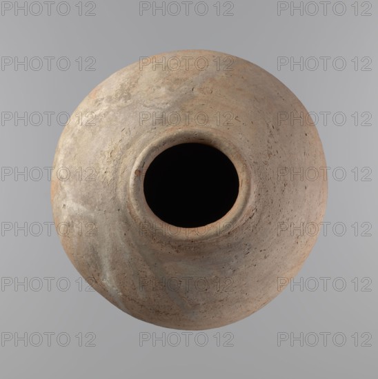 Vessel of an ovoid, slightly downward-stretched shape, widest at about 2/3 of the height. Broad, sloping shoulders, very short, wide neck. Roller-shaped rim, oval in cross-section, slightly flaring. Rounded bottom. NB2 clay according to the Viennese System with a significant amount of organic admixture. The vessel is hand-made from two parts and joined at about 2/3 of the height, turned in the upper part. Surface leveled possibly covered with a layer of engobe. Traces of a white substance in places on the outer surface of the body and on the rim. Vessel colour – beige. Well-preserved vessel. Ca. 2686–2181 BC; Old Kingdom, Edfu; vessel; height 34.6 cm, diameter 11.6 cm rim, diameter 29 cm body., A rounded, weathered clay vessel viewed from above with a dark circular opening at its center, showing a rough, textured surface in shades of tan, beige, light brown, muted gray, and hints of pale green against a smooth gray background.