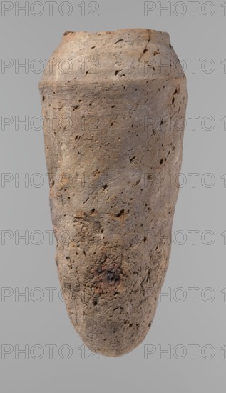 Vessel, Egyptian pottery/ceramic clay hand-made vessel, Old Kingdom c. 2686–c. 2181 BC, from Edfu; vessel height 33.5 cm, rim diameter 10.4 cm, body diameter 15.1 cm. References: form 2., A roughly cylindrical, tapered object with a textured, pitted surface in shades of brown, tan, beige, and hints of reddish-brown, set against a plain gray background; the surface shows irregular indentations, rough patches, and small holes, with areas of darker brown and subtle blackened spots, and the top edge appears uneven and slightly chipped.