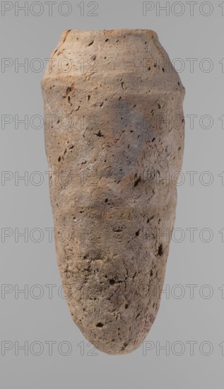 Vessel, Old Kingdom, c. 2686–c. 2181 BC; origin: Edfu; material: clay, hand-made ceramic pottery; form: vessel; height 33.5 cm; rim diameter 10.4 cm; body diameter 15.1 cm., A large, elongated weathered pottery vessel fills the frame, showing a rough, pitted surface with irregular small holes and indentations; the form tapers toward the bottom and has a slightly wider, uneven rim at the top. The colors visible are muted browns, tan, gray, and touches of reddish-brown, with subtle variations and darker grayish areas suggesting surface texture. The background is a smooth, neutral light gray.