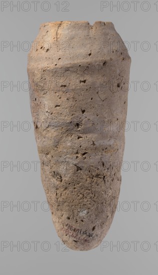 Vessel, Egyptian pottery, hand-made clay vessel; Old Kingdom, c. 2686–c. 2181 BC; from Edfu; vessel, height 33.5 cm, rim diameter 10.4 cm, body diameter 15.1 cm., A tall, weathered clay vessel with a rough, pitted surface occupies the image, showing an elongated, slightly tapered form with an irregular rim that has a small chip. The surface displays a range of earthy colors including light brown, tan, beige, and touches of gray, with darker brown spots and small shadowed cavities across the body. The texture appears coarse and porous with visible pits, cracks, and tiny holes, and there are subtle variations in hue creating faint horizontal banding and mottling. The background is a uniform neutral gray.