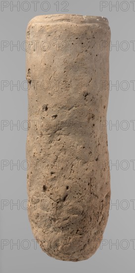 Vessel, Egyptian pottery, ceramics; almost cylindrical hand-made clay vessel, widest just below the rim, rounded bottom, slightly marked narrow sloping shoulders, straight tapering rim. Made of Nile clay with numerous organic inclusions and limestone particles; surface uneven, better worked in upper part, with numerous uneven depressions especially at the bottom. Brown color. Fairly well preserved; surface cracks attributed to careless kneading and firing. Old Kingdom, ca. 2686–2181 BC; from Edfu. Function: vessel. Height 36.5 cm; diameter 11.5 cm rim, 13.2 cm body., A tall cylindrical object with a rough, pitted surface standing against a plain light gray background; the object appears textured and uneven with a weathered, earthy surface showing small holes, indentations, and irregularities; its color is predominantly light brown with areas of darker brown and subtle tan variations; the top edge is slightly rounded and shows a few lighter spots, while the lower portion tapers slightly...
