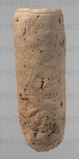 Vessel, Egyptian pottery, handmade clay vessel, almost cylindrical, widest just below the straight tapering rim; rounded bottom; slightly marked, very narrow sloping shoulders. Made of Nile clay with numerous organic inclusions and limestone particles; surface uneven, better worked in upper part, with uneven depressions especially at the bottom. Color: brown. Fairly well preserved; surface cracks resulting from careless craftsmanship in kneading and firing. Old Kingdom, ca. 2686–2181 BC; from Edfu. Vessel; height 36.5 cm, diameter 11.5 cm rim, diameter 13.2 cm body., A vertically oriented cylindrical object with an irregular, textured surface that appears rough and pitted. The object shows earthy tones including shades of brown, tan, beige, and hints of gray. Its top is flattened and slightly uneven, while the sides are rounded with visible small holes, cracks, and indentations. The bottom tapers slightly and also shows rough, crumbly texture with some areas appearing more compacted. T...