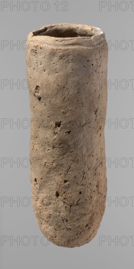 Vessel, Egyptian pottery, handmade clay vessel, almost cylindrical, widest just below the rim, rounded bottom, slightly marked narrow sloping shoulders, straight tapering rim. Handmade of Nile clay with numerous organic inclusions and limestone particles visible on the surface; surface uneven and better worked in the upper part, with numerous uneven depressions especially at the bottom. Color brown. Fairly well preserved; cracks on the surface attributed to careless craftsmanship during kneading and firing. Old Kingdom, ca. 2686–2181 BC; Edfu place of origin. Vessel; height 36.5 cm, diameter 11.5 cm rim, diameter 13.2 cm body., A tall cylindrical object with a rough, pitted surface in shades of beige and light brown, showing irregular dents, holes, and small cracks; the top edge is uneven and slightly thicker with a darker interior, and the background is a smooth neutral gray.