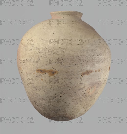 Vessel, Vessel of an ovoid, slightly downward-stretching shape, widest at about 2/3 of the height. Broad, sloping shoulders, very short, wide neck. Rounded rim, slightly profiled in the lower part, oval in cross-section, slightly flaring. Rounded bottom. Just above the widest diameter of the vessel there is a dent made before the vessel was fired. Clay NB2 according to the Vienna System with a significant amount of organic admixture. The vessel was wheel-made, with approximately half of its height worked with a knife or similar tool. The vessel is beige. The vessel is cracked in the upper part. c. 2686–c. 2181 BC; Old Kingdom, Edfu; vessel. Height 34 cm; rim diameter 10.5 cm; body diameter 29 cm., A large rounded pottery vessel with a narrow short neck and flared rim sits against a plain background; the vessel surface is a weathered beige with areas of light tan and subtle pinkish hues, and there are a few rusty brown stains and darker speckles scattered across its textured, slightly s...
