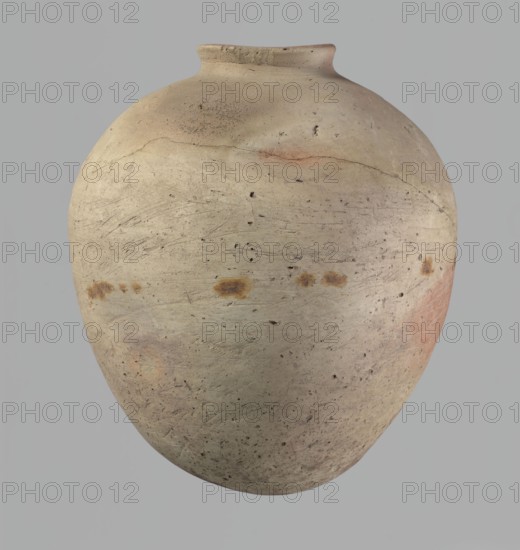 Vessel, Egyptian pottery, clay vessel of ovoid, slightly downward-stretching shape, widest at about 2/3 of the height. Broad, sloping shoulders, very short wide neck. Rounded rim, slightly profiled in the lower part, oval in cross-section, slightly flaring. Rounded bottom. Just above the widest diameter there is a dent made before firing. Clay NB2 Vienna System with significant organic admixture. Wheel-made; approximately half the height was worked with a knife or similar tool. Beige. Cracked in the upper part. Old Kingdom, c. 2686–c. 2181 BC; from Edfu. Vessel; height 34 cm; rim diameter 10.5 cm; body diameter 29 cm., A large rounded ceramic vessel with a narrow neck and flared rim sits against a plain background; the surface shows a weathered, matte finish in shades of beige and light tan with areas of pale gray, faint orange and rusty brown spots, and subtle pinkish patches, along with numerous small dark specks, shallow scratches, and hairline cracks across the body.