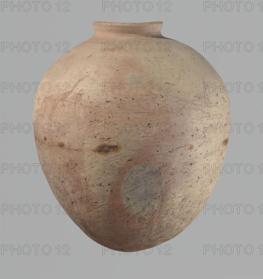 Vessel, Egyptian pottery, ceramics, clay vessel. Ovoid, slightly downward-stretching shape, widest at about 2/3 of the height; broad, sloping shoulders; very short, wide neck; rounded rim, slightly profiled in the lower part, oval in cross-section, slightly flaring; rounded bottom. Dent just above the widest diameter made before firing. Clay NB2 Vienna System with significant organic admixture. Wheel-made; approximately half the height worked with a knife or similar tool. Beige. Cracked in the upper part. Old Kingdom, c. 2686–c. 2181 BC; Edfu. Vessel; height 34 cm; rim diameter 10.5 cm; body diameter 29 cm., A large rounded ceramic vessel with a narrow neck and flared rim shown against a plain background; the surface is textured with visible scratches, pits, and weathered areas, and shows a palette of pale beige, light tan, muted pink, and soft gray with a few darker brown spots and smudges.