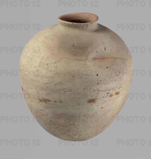 Vessel, Egyptian pottery, ceramics, clay vessel. Ovoid, slightly downward-stretching shape, widest at about two-thirds of the height; broad, sloping shoulders; very short, wide neck; rounded rim, slightly profiled in the lower part, oval in cross-section, slightly flaring; rounded bottom. Dent just above the widest diameter made before firing. Clay NB2 Vienna System with significant organic admixture. Wheel-made; approximately half the height worked with a knife or similar tool. Beige. Cracked in the upper part. Old Kingdom, c. 2686–c. 2181 BC; from Edfu. Vessel; height 34 cm; rim diameter 10.5 cm; body diameter 29 cm., A rounded clay vessel with a narrow neck and flared rim, showing a textured, uneven surface with a few rough patches and small pits; the body displays subtle horizontal tool marks and faint surface cracks, along with several darker spots and smudges. The visible colors include light beige, pale tan, muted brown, touches of warm reddish-brown, and small areas of darker b...
