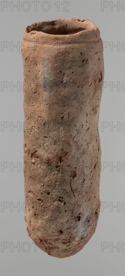 Vessel, Egyptian pottery, handmade clay vessel, almost cylindrical, widest just below a straight, slightly tapering rim; rounded bottom; shoulders almost illegible. Handmade from Nile clay with numerous organic inclusions and limestone particles visible on the surface; carelessly made with uneven depressions especially at the bottom; brown color; cracks from clay preparation and firing. Date ca. 2686–ca. 2181 BC Old Kingdom, from Edfu. Vessel, height 37.9 cm; diameter 11.4 cm rim, 12.5 cm body., A tall cylindrical clay vessel with an irregular rim and rough, pitted surface, showing a weathered texture and small holes and indentations across its body; the form is slightly tapered with subtle bulges and a rounded base, and visible marks and scratches on the exterior. The colors visible are various shades of brown, reddish-brown, gray, and a few darker brown spots, set against a neutral light gray background.